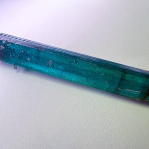 May include: A raw, elongated crystal with a striking teal and blue-green color. The gemstone has a rough, textured surface and a natural, unpolished appearance. The crystal is a vibrant example of natural mineral formations.
