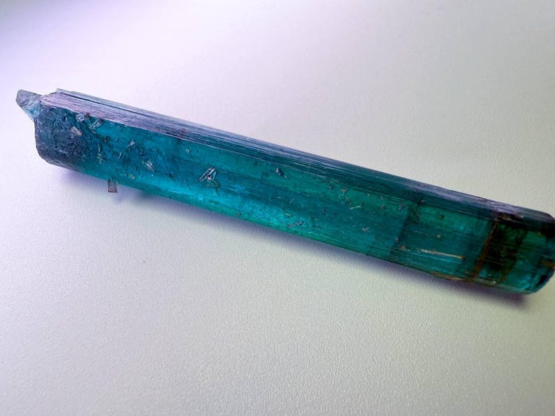 May include: A raw, elongated crystal with a striking teal and blue-green color. The gemstone has a rough, textured surface and a natural, unpolished appearance. The crystal is a vibrant example of natural mineral formations.