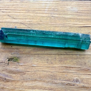 May include: A raw, elongated teal tourmaline crystal with a dark blue tip rests on a weathered wooden surface. The crystal's facets reflect light, showcasing its translucent quality. The wood grain adds a natural, rustic contrast to the gemstone.