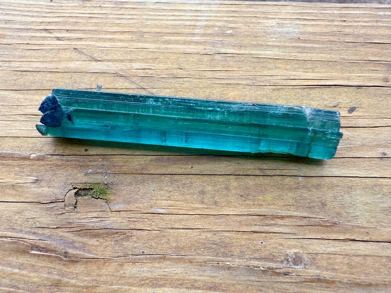 May include: A raw, elongated teal tourmaline crystal with a dark blue tip rests on a weathered wooden surface. The crystal's facets reflect light, showcasing its translucent quality. The wood grain adds a natural, rustic contrast to the gemstone.