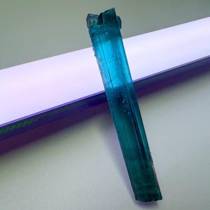 May include: A vibrant, elongated blue-green tourmaline crystal. The gemstone is a striking shade of teal, with visible striations and a natural, rough texture. The crystal is positioned diagonally against a white and purple background.