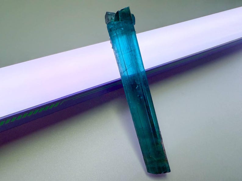 May include: A vibrant, elongated blue-green tourmaline crystal. The gemstone is a striking shade of teal, with visible striations and a natural, rough texture. The crystal is positioned diagonally against a white and purple background.