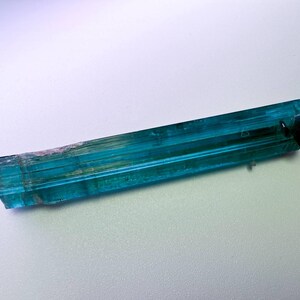 May include: A vibrant, elongated teal tourmaline crystal with a dark, fractured end. The gemstone displays a translucent quality, with visible striations and subtle color variations. The crystal is approximately 3 inches long.