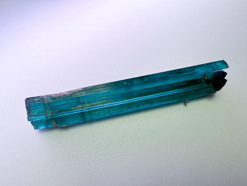 May include: A vibrant, elongated teal tourmaline crystal with a dark, fractured end. The gemstone displays a translucent quality, with visible striations and subtle color variations. The crystal is approximately 3 inches long.