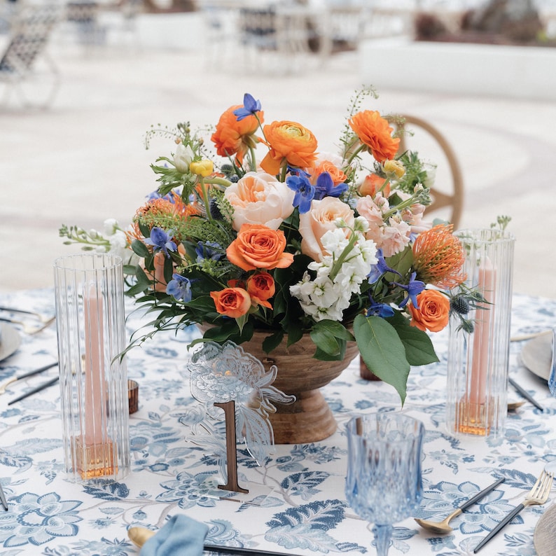 May include: A table setting with a floral centerpiece featuring orange roses, blue delphiniums, and white flowers in a wooden bowl. The table is covered with a blue and white floral tablecloth, with clear glass candle holders and a table number.