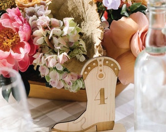 May include: A wooden table number in the shape of a cowboy boot, with the number 4 etched on the front. The boot is light brown with decorative stitching and star accents. The boot sits on a rectangular base. A floral arrangement is in the background.