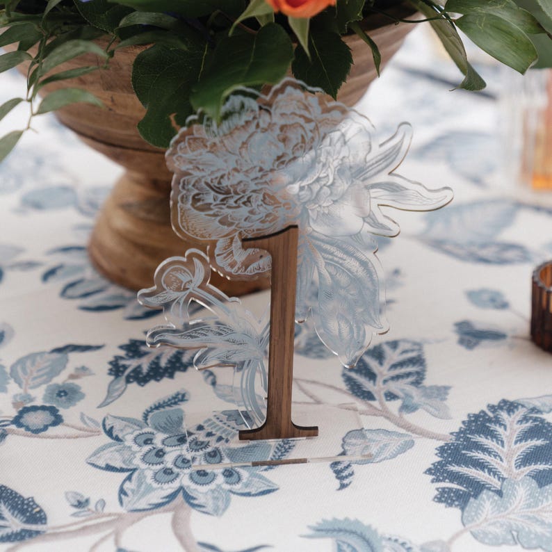 May include: A clear acrylic table number one with a floral design, set on a wooden base. The number is displayed on a table with a blue and white floral tablecloth. A wooden bowl with flowers is in the background.