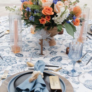May include: A table setting with a floral centerpiece, candles, and place settings. The table is covered with a blue and white floral tablecloth. A clear acrylic table number with the number 1 is visible. The setting includes blue glassware, gold flatware, and a blue napkin.