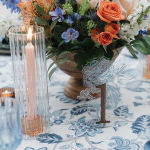 May include: A table setting with a floral centerpiece, a lit pink candle in a clear glass holder, and a table number. The table number is a clear acrylic flower design with the number "1" in brown. The tablecloth has a blue floral pattern.