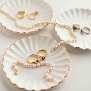May include: Three white ceramic dishes with scalloped edges and gold trim, each holding a pearl necklace and rings. The dishes are arranged on a white surface.