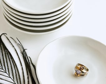 May include: A stack of six small, white ceramic plates with a slightly raised rim. One plate holds a gold ring with two small gemstones. A dark, feathery accent is in the lower left corner.