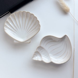 May include: Two white ceramic dishes shaped like seashells. The dishes are on a white surface.
