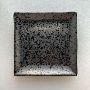 May include: A black square ceramic plate with a speckled finish. The plate has a slightly raised rim.