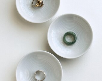 May include: Three small, white ceramic ring dishes with a textured, concentric circle design. Each dish holds a ring: a gold ring with a gemstone, a green ring, and a silver ring. The dishes are arranged on a white surface.