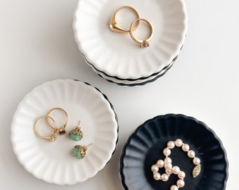 Peut inclure: Trois petits plats décoratifs en céramique avec des bords festonnés. Deux plats sont blancs avec des rayures noires et un est noir. Les plats sont remplis de bijoux, dont des bagues, des boucles d'oreilles et un collier de perles.