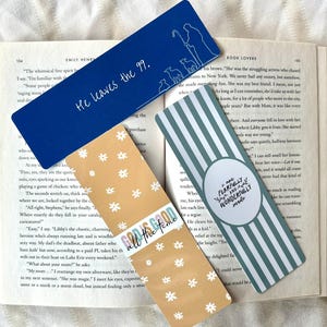May include: Four decorative bookmarks rest on an open book. One is blue with white text and a shepherd illustration. Another is tan with white daisy patterns and text. The third is striped with text.