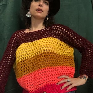Peut inclure: Un haut en crochet multicolore avec un motif rayé dans les tons marron, jaune, orange, rose et rouge. Le haut est ample et a un col large.