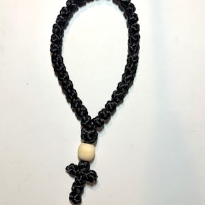May include: Black knotted prayer rope with a wooden cross and bead. The rope is made of tightly woven black threads, forming a loop. The cross and bead are light-colored wood. The background is white.