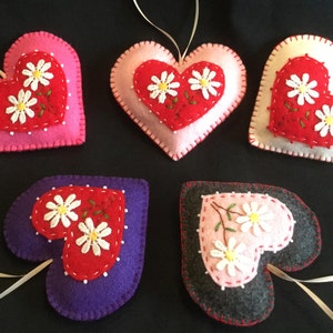 May include: Five felt heart ornaments with embroidered white daisies and green stems. The hearts are in various colors: pink, red, white, purple, and gray.