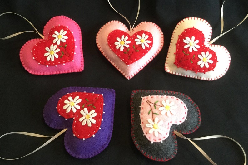 May include: Five felt heart ornaments with embroidered white daisies and green stems. The hearts are in various colors: pink, red, white, purple, and gray.