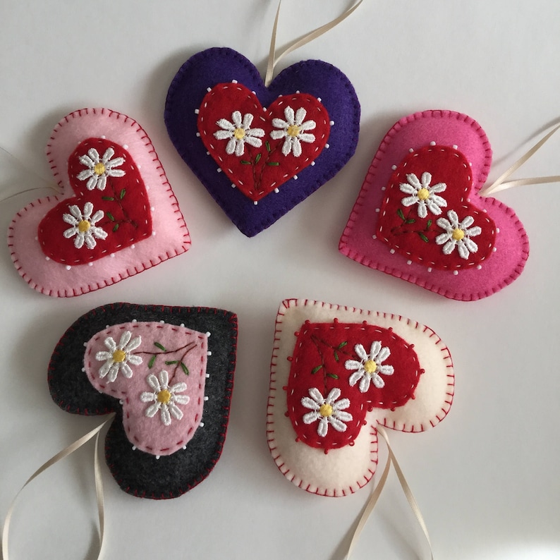 May include: Five felt heart ornaments with embroidered daisy flowers. The hearts are in shades of pink, red, white, purple, and gray.