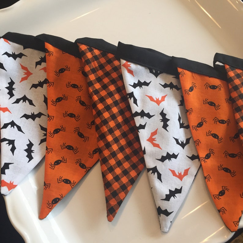 May include: A Halloween banner with six triangular flags. The flags are made of fabric and feature different patterns, including bats, spiders, and a black and orange plaid. The flags are sewn together with black thread.