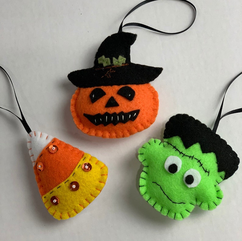 May include: Three felt Halloween ornaments: a pumpkin with a black witch hat, a green Frankenstein, and an orange and yellow candy corn.