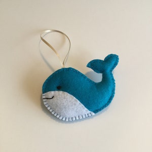 May include: A teal blue felt whale ornament with a white felt belly and a black eye. The whale has a stitched smile and a white ribbon for hanging.