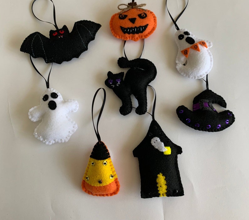 May include: A set of felt Halloween ornaments, including a black bat, a black cat, a pumpkin, a witch's hat, a ghost, a candy corn, and a black house with a ghost inside.