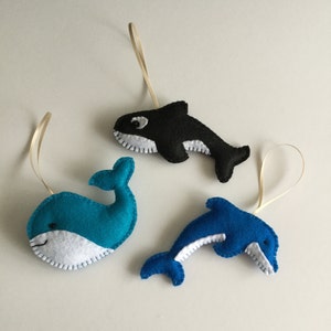 May include: Three felt ornaments shaped like a whale, a dolphin, and an orca. The whale is blue with a white belly, the dolphin is blue with a white belly, and the orca is black with a white belly.