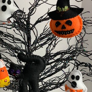 May include: A black felt cat, a white felt ghost with an orange and white "BOO" banner, a felt pumpkin with a black witch hat, and a felt candy corn ornament hanging on a black twig tree.
