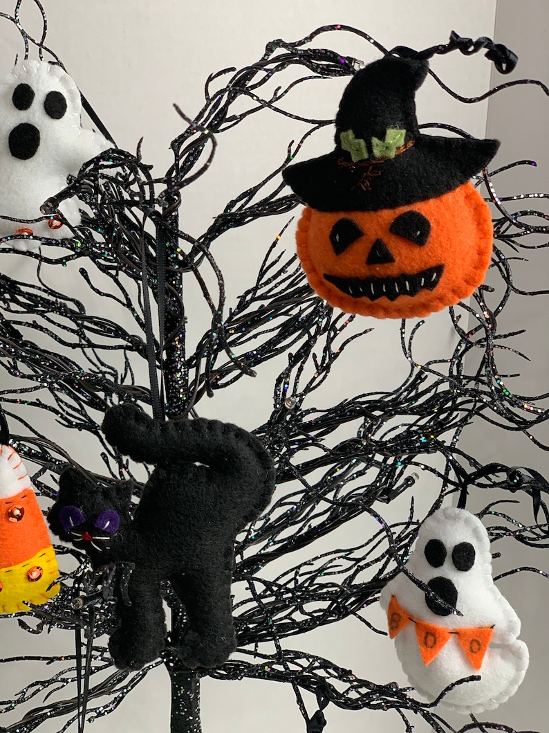 May include: A black felt cat, a white felt ghost with an orange and white "BOO" banner, a felt pumpkin with a black witch hat, and a felt candy corn ornament hanging on a black twig tree.