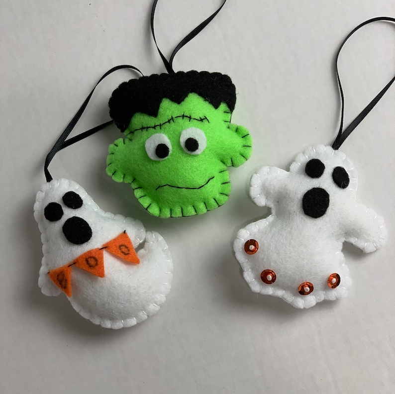 May include: Three felt Halloween ornaments: a green Frankenstein, a white ghost with orange bunting, and a white ghost with black eyes and orange sequins.