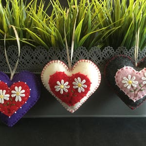 May include: Three felt heart ornaments with embroidered white daisies. The hearts are purple, red and white, and gray and pink.