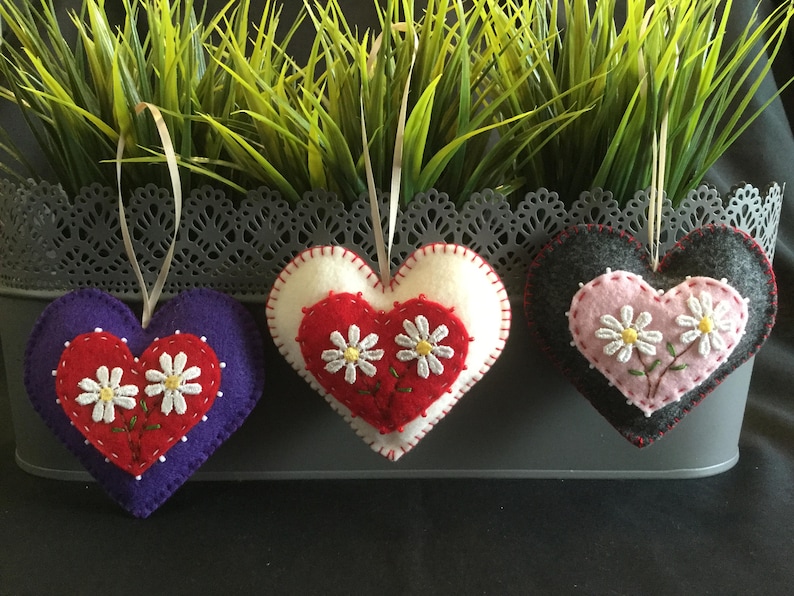 May include: Three felt heart ornaments with embroidered white daisies. The hearts are purple, red and white, and gray and pink.