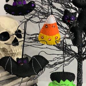 May include: Five felt Halloween ornaments hanging on a black twig tree. The ornaments are a black witch hat, a black bat, a green Frankenstein, a black cat, and an orange and yellow candy corn.