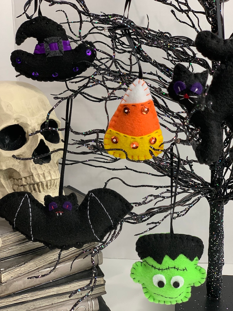May include: Five felt Halloween ornaments hanging on a black twig tree. The ornaments are a black witch hat, a black bat, a green Frankenstein, a black cat, and an orange and yellow candy corn.