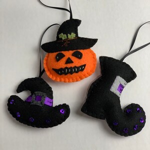May include: Three felt Halloween ornaments: a jack-o'-lantern with a black hat, a black witch's hat with a purple ribbon, and a black witch's boot with purple sequins.