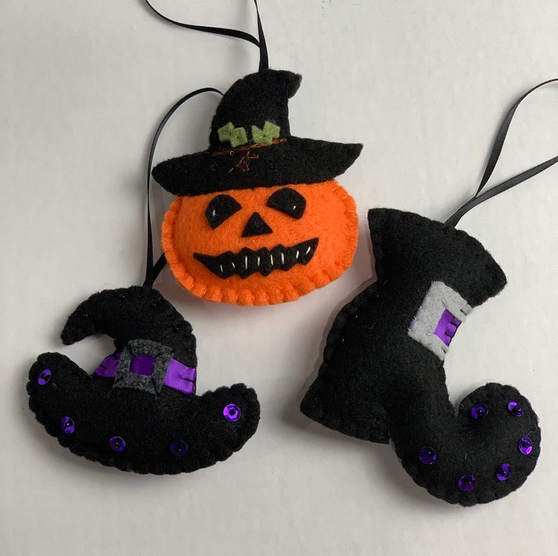 May include: Three felt Halloween ornaments: a jack-o'-lantern with a black hat, a black witch's hat with a purple ribbon, and a black witch's boot with purple sequins.