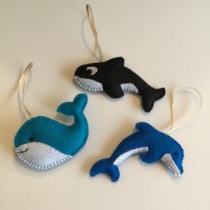May include: Three felt animal ornaments: a blue whale, a blue dolphin, and a black and white orca. Each ornament has a white felt belly and a white felt trim around the edges. The ornaments are hanging from white ribbon loops.