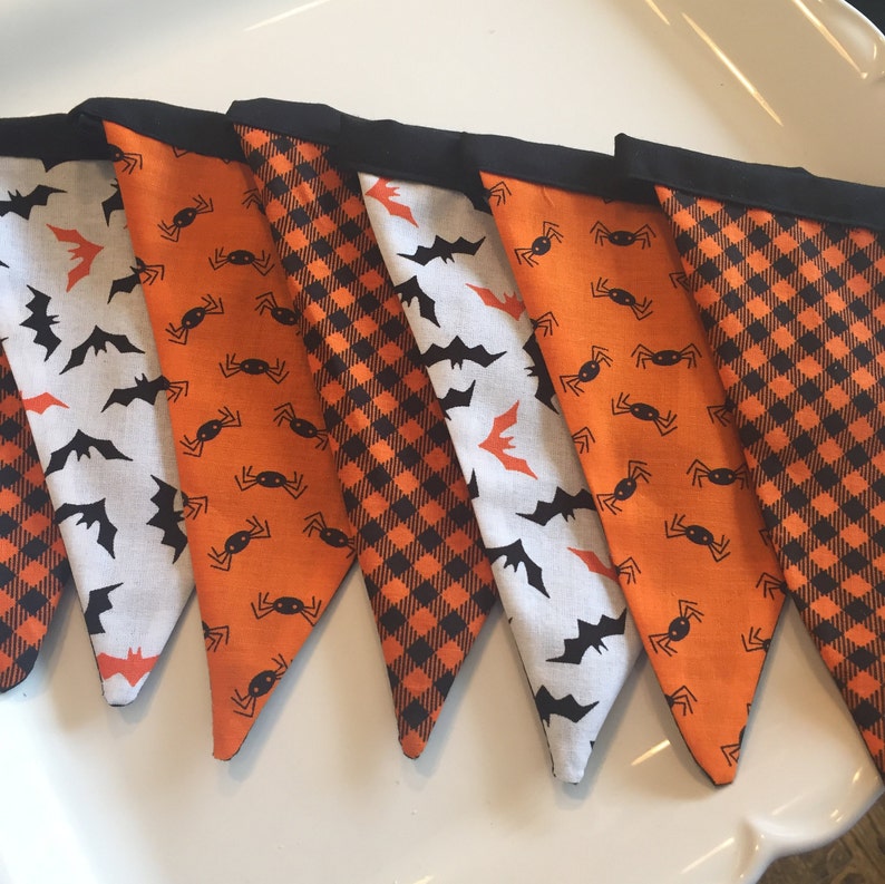 May include: A Halloween banner with six triangular flags. The flags are made of orange and black fabric with different patterns. One flag has a black and orange plaid pattern, another has a white background with black bats, and the rest have an orange background with black spiders.