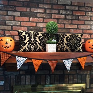 May include: A brick fireplace mantel decorated for Halloween with a black and gold patterned pillow, a small potted tree, and a banner of orange, black, and white pennants with bats and checkered patterns. Two orange jack-o'-lantern buckets are on either side of the mantel.