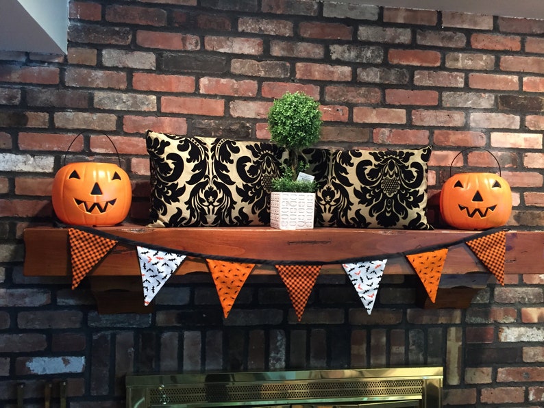 May include: A brick fireplace mantel decorated for Halloween with a black and gold patterned pillow, a small potted tree, and a banner of orange, black, and white pennants with bats and checkered patterns. Two orange jack-o'-lantern buckets are on either side of the mantel.