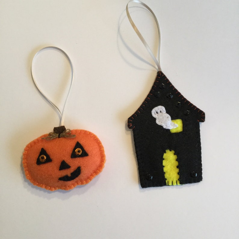 May include: Two felt Halloween ornaments, a pumpkin and a house. The pumpkin is orange with a black face and gold glitter eyes. The house is black with a yellow door and a white ghost inside.