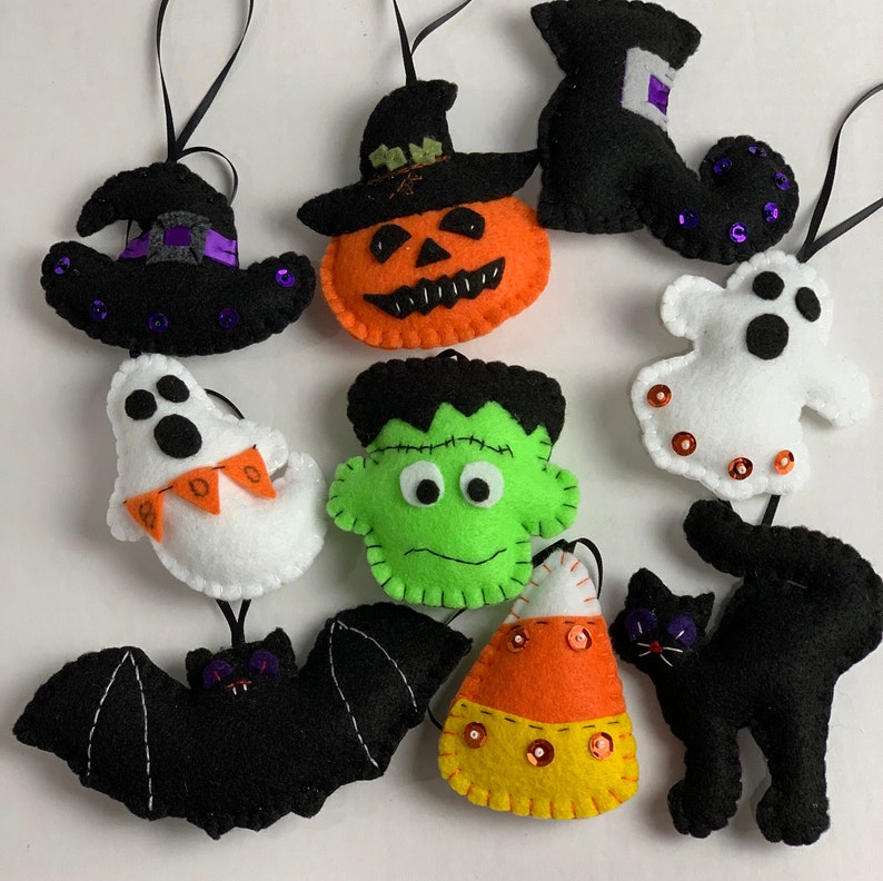 May include: Nine felt Halloween ornaments with various designs, including a pumpkin, a ghost, a witch's hat, a bat, a Frankenstein, a candy corn, and a black cat. Each ornament is decorated with sequins and has a loop for hanging.