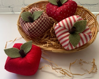 May include: Four red fabric apples in a wicker basket. The apples are decorated with different patterns, including gingham, polka dots, and stripes. The apples have green felt leaves and brown stems.