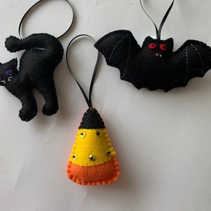 May include: Three felt Halloween ornaments: a black cat, a black bat with red eyes, and a yellow and orange candy corn.