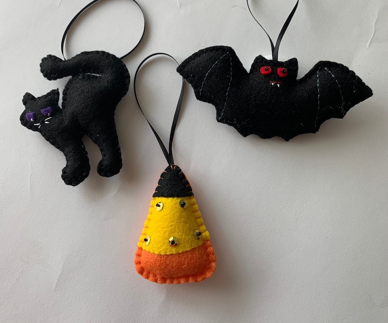 May include: Three felt Halloween ornaments: a black cat, a black bat with red eyes, and a yellow and orange candy corn.
