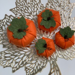 May include: Four felt pumpkins in various sizes, with green felt leaves and stems, arranged on a gold leaf-shaped placemat.
