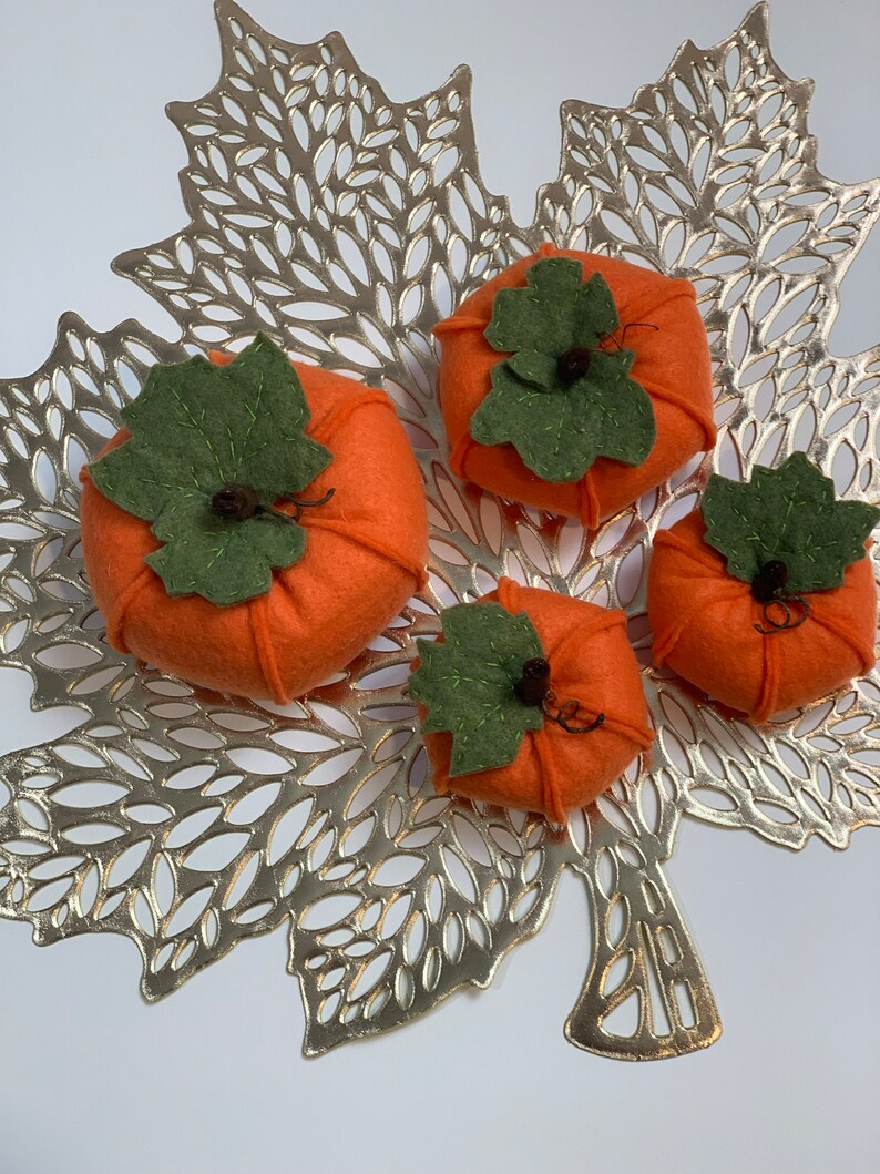 May include: Four felt pumpkins in various sizes, with green felt leaves and stems, arranged on a gold leaf-shaped placemat.